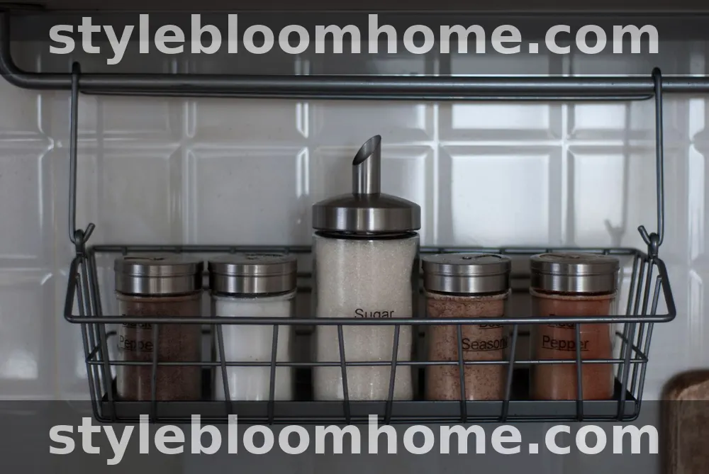 A hanging wire basket mounted on a kitchen wall rail, holding five labelled glass jars of pepper, seasoning, sugar, and salt against white tiles